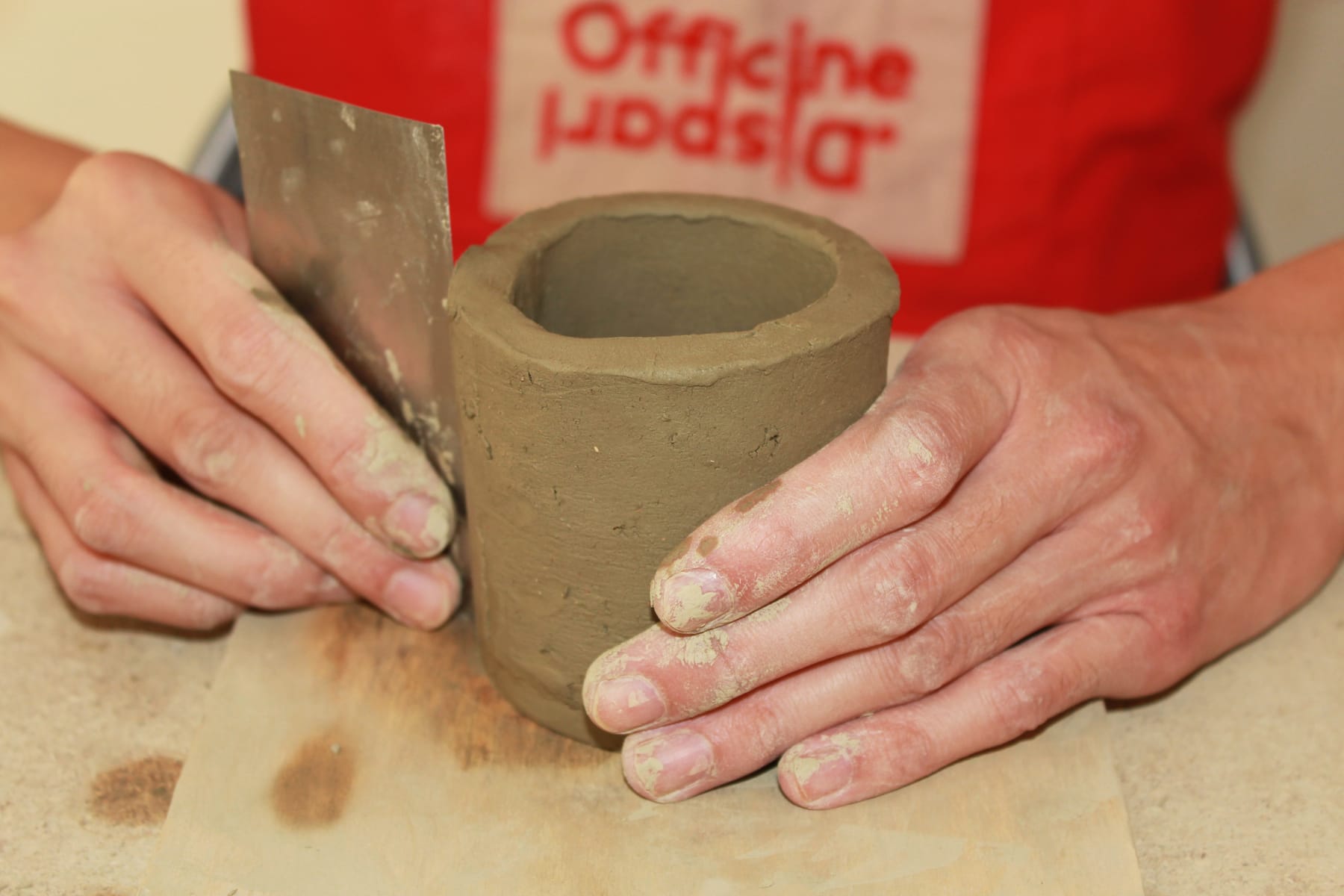 Mani di un artigiano sporche di creta che modellano un vaso cilindrico grezzo utilizzando una spatola di metallo su un piano di legno.